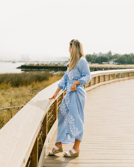 Model walking along a curved coastal boardwalk wearing white linen ruffle two-strap espadrille wedge sandals with a blue embroidered linen maxi wrap dress, overlooking marsh grass and harbor | White Linen Ruffle