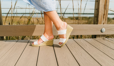Side view of white linen ruffle two-strap espadrille wedge sandal with scalloped edges and jute-wrapped heel, worn with a light blue linen dress on a coastal boardwalk | White Linen Ruffle